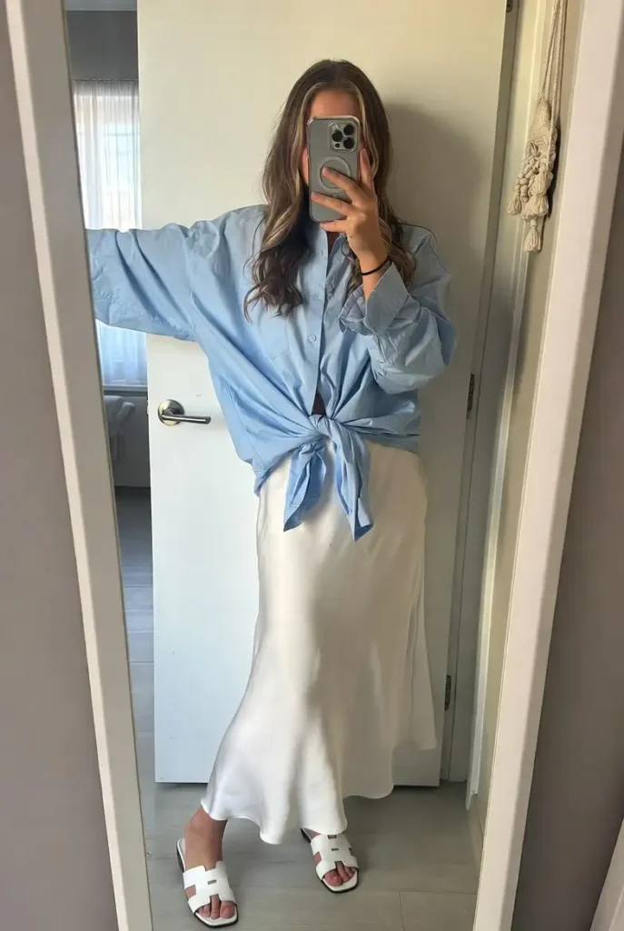 One of the 13 White Maxi Skirt Outfits: a light blue oversized shirt tied at the waist over a white satin maxi skirt.