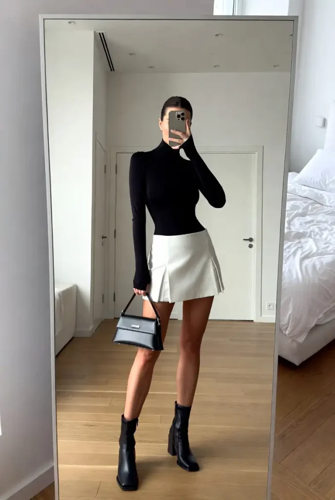 "White short skirt outfit idea paired with a fitted black turtleneck, black mini bag, and ankle boots"