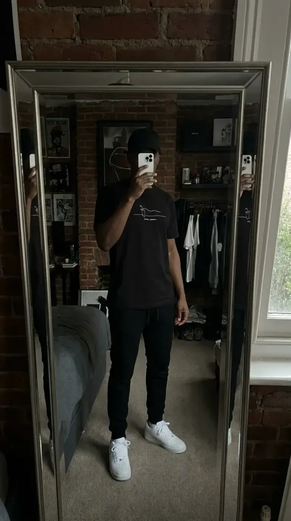 Casual black men outfit ideas: graphic black tee, slim joggers, black cap, and white Air Force 1 sneakers.