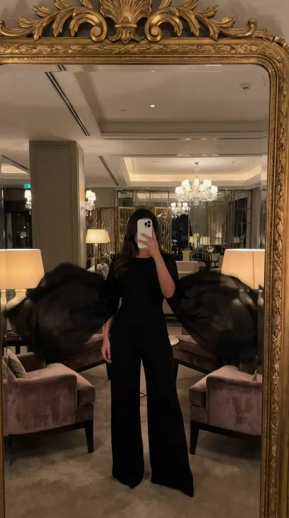 Elegant black classy wedding guest jumpsuit summer featuring dramatic sheer cape sleeves.

