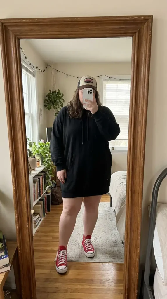 Red and black outfit ideas for plus size: black hoodie mini dress paired with red Converse sneakers and a trucker hat.
