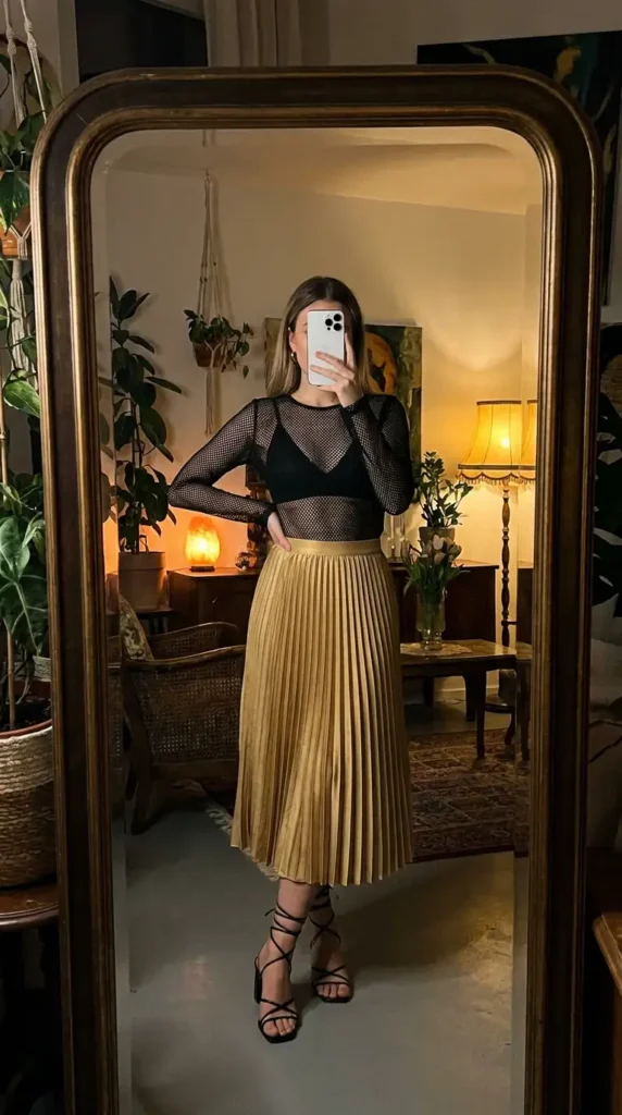 Casual black and gold party outfit ideas with a sheer black mesh top paired with a pleated gold satin midi skirt.
