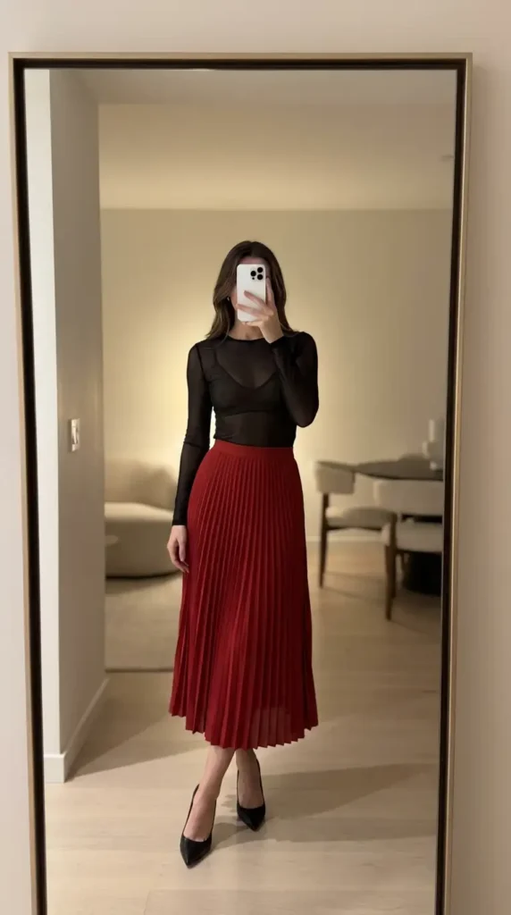 Black sheer long-sleeve top layered over a red pleated midi skirt, a stunning red and black outfit idea for party.
