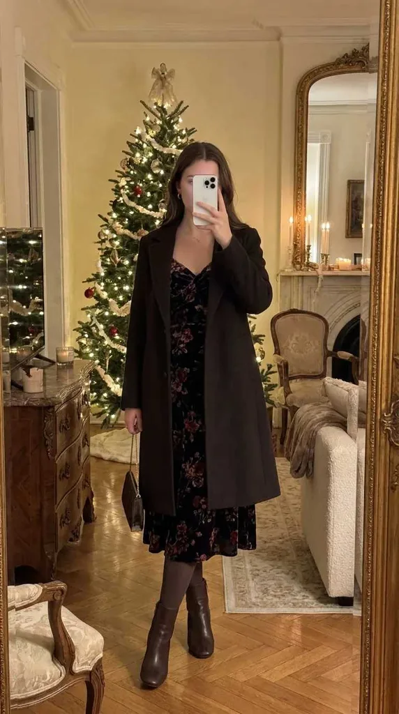 Woman in dark floral midi dress, black coat, boots and bag posing by Christmas tree for New Year Outfit Ideas Parties.
